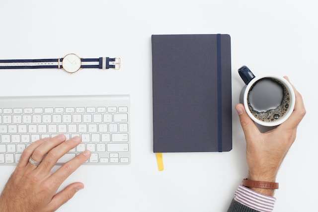 A man's left hand is typing on a white key board, looks like it is an apple keyboard. Above the keyboard a gold rimmed watch with white face and canvas strap lay on the table. The strap is a white strip pattern in the middle of a navy blue canvas base with matching golden buckles. The man's right hand is holding a mug of black coffee. This picture is shot from above only showing the man's two hands, keyboard, coffee mug and a dark purple cover note book sits in between the man's two hands, and on the right side of the keyboard. The notebook has a yellow cloth bookmark sticking out from the bottom of it. A navy blue elastic band close the notebook neatly. Maybe he is working on one of the the most influential Coffee blog websites.