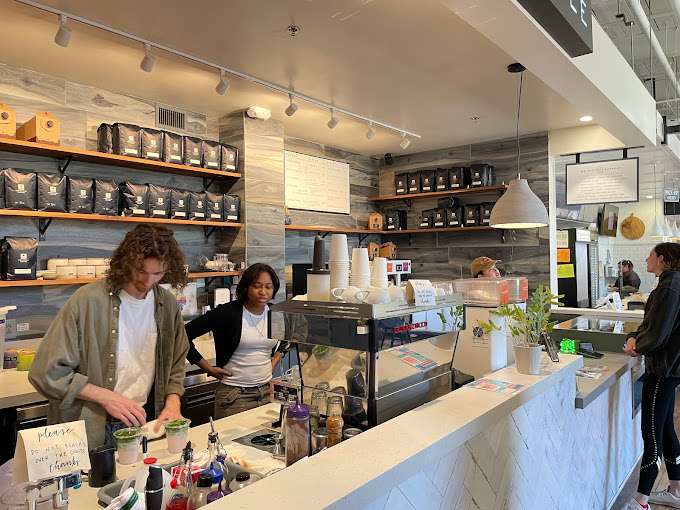 The serving counter inside Undercurrent Coffee. This photo was shot at an angle, a male barista in the foreground waring a green long sleeve button shirt without button it up showing the white t-shirt underneath. He is preparing two pink drinks with green tops, they might be some kind of green tea drinks. A black woman next to him looks at the man's hands. The interior design of this shop is very modern and sleek with hard sharp edge counter with with white and dark grey counter tops at different level, gives a pleasant visual contrast while you are looking at the counter.