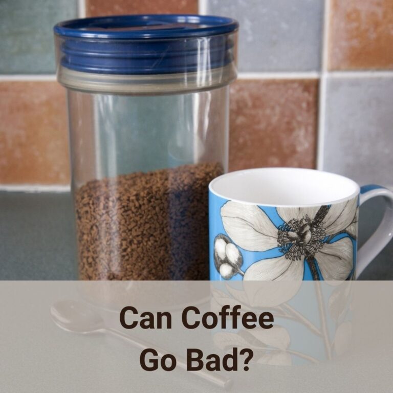 On a dark gray kitchen counter, a round cylinder shape clear plastic food container has instant coffee crystals in it. The mosaic back splash tiles are in the background. A coffee mug is at the front of the food container slightly towards the right. The white ceramic coffee mug external has a sky blue glaze as the backgound, in the foreground are large black-and-white line art of magnolia flowers, flower buds, broad leaves and branches. Can coffee go bad? Yes, it can!