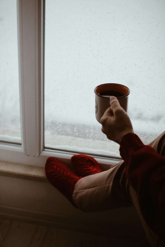 A woman holding a cup of coffee with her left hand and sitting next to a glass door appreciating the white snow in the yard outside through the glass. Coffee and snow are the two usual elements in Nordic people's life.