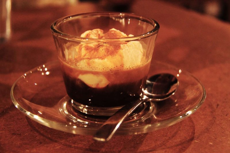 Affogato, a shot of espresso poured over a scop of ice-cream, in a short glass with a saucer and a stainless steel desert spoon next to the cup.