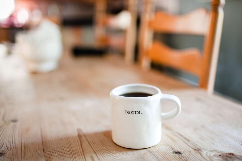 A white mug of black coffee on a long wooden table. Black letters of "Begin" are engraved on the white mug. Coffee has positive impact on people's mood. This is one of the best benefits that coffee boosts your performance.