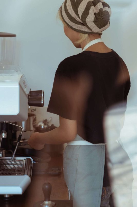 a blonde barista, with a black short sleeve t-shirt and a grey apron that has white neck and waist straps, is making coffee using a coffee machine, but blonde espresso is nothing to do with a blonde making coffee, it is a type of light roast coffee is pulled as espresso. Blonde espresso is stronger than regular espresso in term of caffeine content.