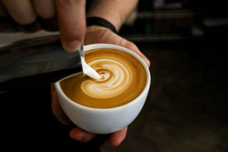 A barista pouring cream into a small cup of cafe latte which is a milky hot coffee.