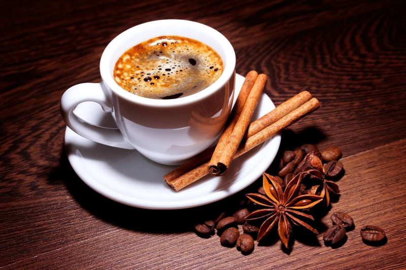 A white ceramic cup of black coffee with matching saucer on a dark brown wooden table top. The coffee has a layer of crema and two sticks of cinnamon in the saucer for sweetening coffee naturally. Some coffee beans and two clove of star anise loosely lay next to the saucer.