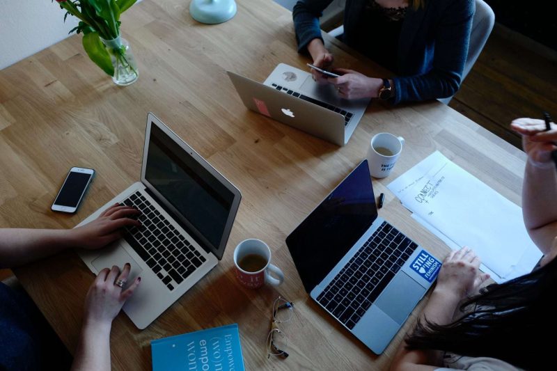 Three people with their laptops are working at a big rectangular table, two women have two mugs of coffee on the table next to their laptops. Coffee increases alertness and focus, which is one of the benefits that coffee boosts your performance at work.