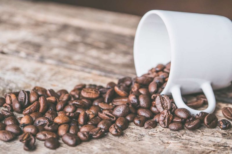 On a rustic wooden table top, a white ceramic coffee mug lays on its side and coffee beans spill out of the mug onto the table. The mug is on the right side of the photo , the coffee beans spill towards the left side in the photo. Is coffee a drug?