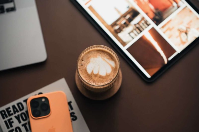 This photo is shot from above. A glass of mocha with white cream leafy pattern sits in the center of a table top, a corner of a silver laptop can be seen on the upper left side of the coffee glass. A book on the right upper corner of the picture is out of focus, so it is difficult to tell what these pictures on the cover are about. The lower right corner is showing a corner of a magazine, to top of the magazine is a cell phone with an orange protection case. This cell phone has 3 camera lenses, maybe it is an iPhone. These are the basic equipment for running one of most influential coffee blog websites.