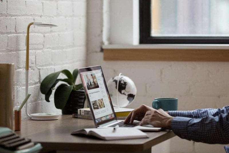 A man is sitting at a desk which its right side against a window. On the desk, there is a laptop that the man is working on. There is a pot of orchid at the right corner of the desk and a study lamp on the left side of the plant pot. Only the man's two arms and hands are visible in the photo, he wears a blue and white plaid long sleeve shirt. Both of his hands are on the keyboard of the laptop as if he is typing away some articles. On the right side of the man's right hand a teal color coffee mug sits on the desk. An opened notebook is on the left side of the man's left hand. A pen, out of focus of the lens, is sitting on top of the open notebook. He is working on the most influential coffee blog website.