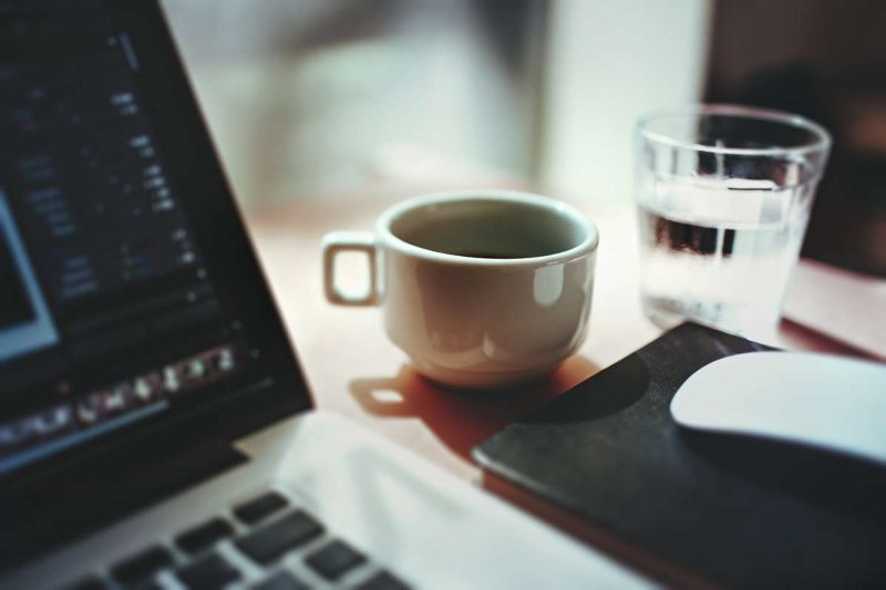 In the center of this photo is a coffee cup, on its right a glass of water is sitting there. A corner of a laptop is shown in the picture on the 1/3 of this photo's left side. A partial of the black screen border and the screen background can be seen here. The keyboard has silver edge with black key buttons. On the right to the laptop there is a rectangular hard surface, maybe a iPad of something similar just in front of the coffee cup. On top of this reflective hard surface, maybe the pad's screen, there is a white computer mouse sits there. 2/3 of the mouse can be seen in this picture. A laptop and a cup of coffee that is all you need to start a most influential coffee blog website.