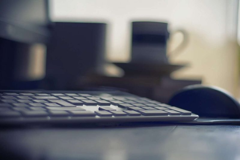 The foreground's black keyboard is very clear, the background of this picture is out of focus, so everything is blurry. A few thing still can be made out by their silhouette. A computer mouse is on the right side to the keyboard. A mug with a saucer is on a counter top, but it is blurry, it is really hard to make a definite description.