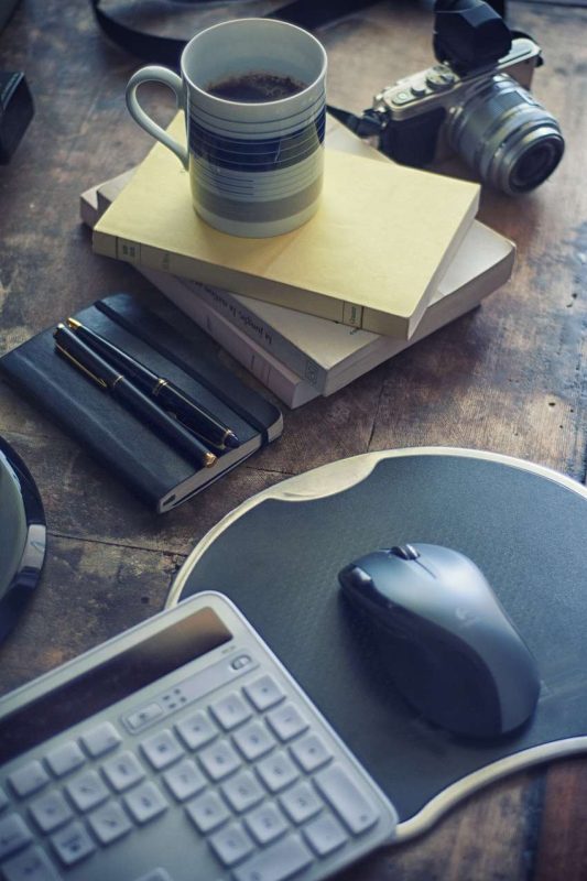 A mug of black coffee sits on top of stack of books, actually only three books. These objects are all sitting on a rustic, maybe even recycled timber table top. To the right of this stack of 3 books is a nice camera with a flash light mounting on top of it. The lens cap is off. The camera has a black neck strap laid loosely on the table. To the left side of the book stack a navy blue cover notebook with black elastic closure strap sits there. Two pens are on top this notebook, one is black body the other is in navy blue body. Both pens have golden highlight parts visible to give them a more classy look. Below the notebook a round mousepad sits on the tap, on it is a black computer mouse. 1/5, left part of the mousepad is covered by a keyboard, only the number keys of this keyboard is visible in the picture. Good pictures and great notes are key element for writing a most influential coffee blog website.