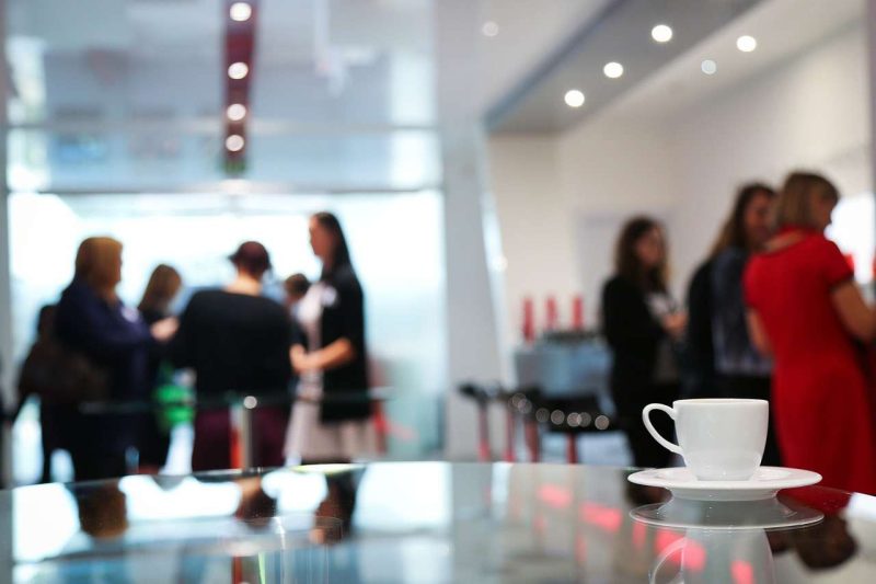 In a well lit corporate coffee break room women gather in two groups chatting in the background, they are out of focus. In the foreground is a round coffee table with a white ceramic coffee cup with matching saucer on it, the table surface is very flat and reflective, so the reflection of the coffee cup saucer set is clearly visible besides the reflection of the two group woman's heads too. Coffee breaks promote collaboration and social interaction, which is one of the benefits that coffee boosts your performance at work.