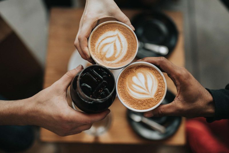 The picture is taken from the top. Three hands holds a cup of coffee each and in the middle of clinking glass. The three coffee clink together forming an symmetrical triangle. The right and the top cups are creamy coffee in ceramic cups. The left lower one is in a glass and it is a black coffee with some ice in it. A square wooden coffee table is in the background but it is quite blurry. You still can make out that there are two black saucer with silver teaspoons sit on the table. Social gathering in a coffee shop is very common nowadays as part of modern life style. Is coffee a drug?