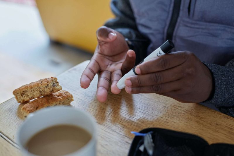 Sitting at a table, a black man is using a marker-pen-shape-like blood glucose meter to poke his right ring finger tip. A cup of coffee and two pieces of biscotti are on the table in front of the man's hands.