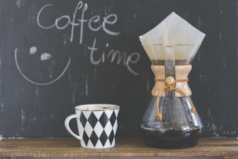 This picture shows a narrow wooden bench is against a blackboard. On the the bench is a drip coffee maker on the right and a white porcelain mug with black diamonds patten on the left, next to the drip coffee maker. The mug has a golden rim on the top and the coffee is near the edge of the mug. A wet filter cone is on the drip coffee maker and the translucent filter shows the coffee ground in it. The the black board just above the white mug, "Coffee time" with a smile face is scribbled on in white chalk. Is coffee good for you? It depends.