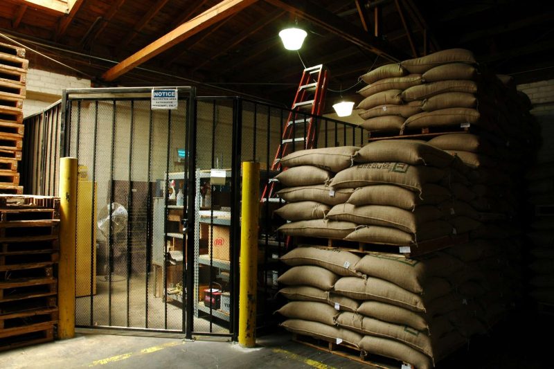 This picture shows a corner of a coffee bean warehouse. On the right of the picture there are stacks of sacks of raw coffee beans in the factory lights shadow. On the left an area is locked with a meshed fance and door. Inside this locked space there are some shelves for storage, it may be for cleaning stuff which are shown and two car batteries at the bottom of one of the shelves. Two tall round steel bollard posts flank the pad locked mesh steel gate. They are painted in bright yellow paint for warehouse safety operation. Coffee has changed the world through it trade which started at least 500 years ago in large scale.