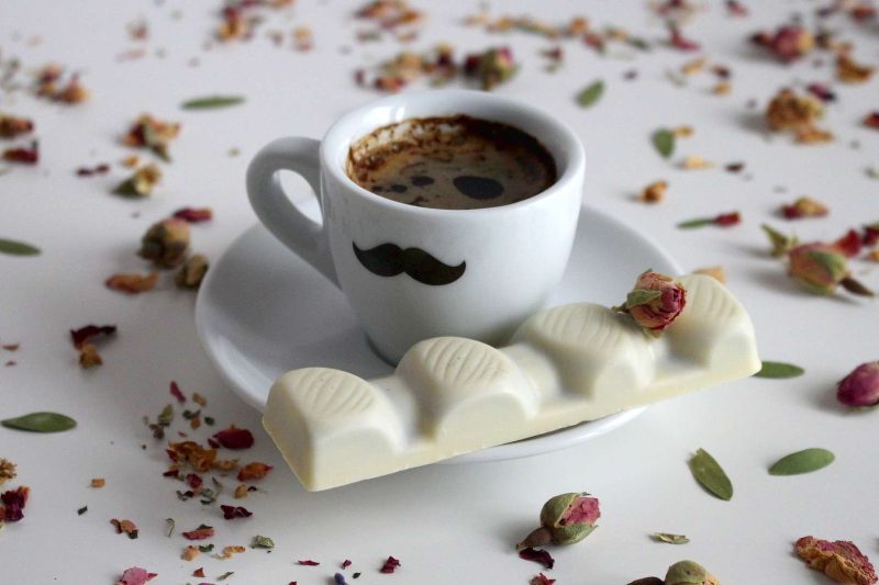 A cup of mocha with matching white saucer on a table top. The table top is lit with dry wild roses. A piece of white chocolate is on the saucer next to the cup. an "easy mocha coffee" recipe primarily lies in the ingredients and complexity of the preparation.