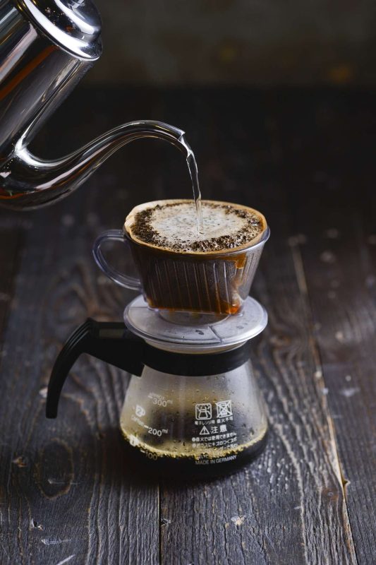 Hot water, in a stainless steel pot, is poured over a glass drip brew coffee pot that is on a dark color timber table top.