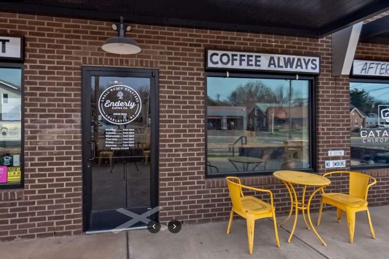 Front Door of Enderly Coffee Co, which is one of the most popular coffee shops in charlotte nc. The photo show a yellow four leg metal round coffee table with two matching color metal chairs; they are to the right of the front glass door in front of a large shop window. A large painted black overhanging over the shop front provides some weather covering for people like to sit outside the coffee shop to enjoy their cup of joe.