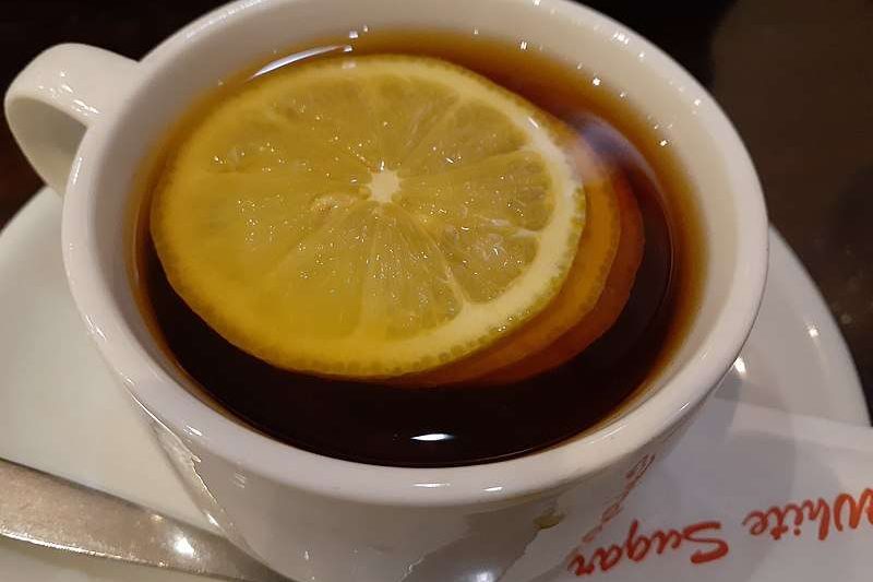 3 pieces of lemon in a white cup of black coffee with matching white saucer and a stainless steel tea spoon next to the cup, this is a type of espresso romano.