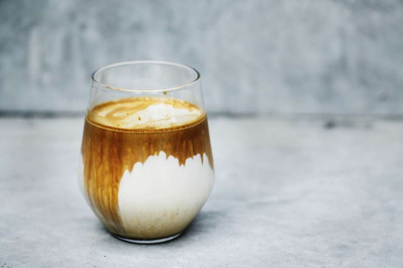 A glass tumbler of very creamy coffee sits on a table with a blurry background. The cream is more than the coffee, so the lower part in the tumbler, it is white which means the cream replaced the coffee at the bottom. Is coffee good for you? If you drink coffee with too much additives, it may not be that good.