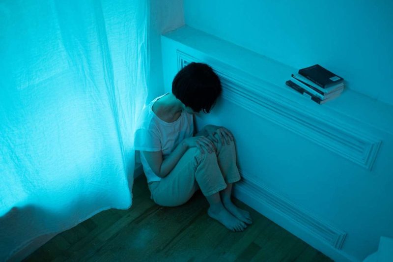 A woman is sitting on the hardwood floor at the corner next to a window. The blueish dim light implies the setting is in a night. A white curtain covers the window. The breeze from outside seems to disturb the white curtain's right lower corner where the woman sits. She sits there in a fetal position has her head lowered toward her two hands which are on her knee. Her left side of the head is resting on a the panel of a narrow ledger. This photo shooting angle is above the woman, so her face is blocked by her short dark hair. She might be an Asian woman and suffering from insomnia, in some kind of pain or sadness. A stack of books are on the narrow ledger which height is just above this woman's head. Is coffee good for you? It depends.