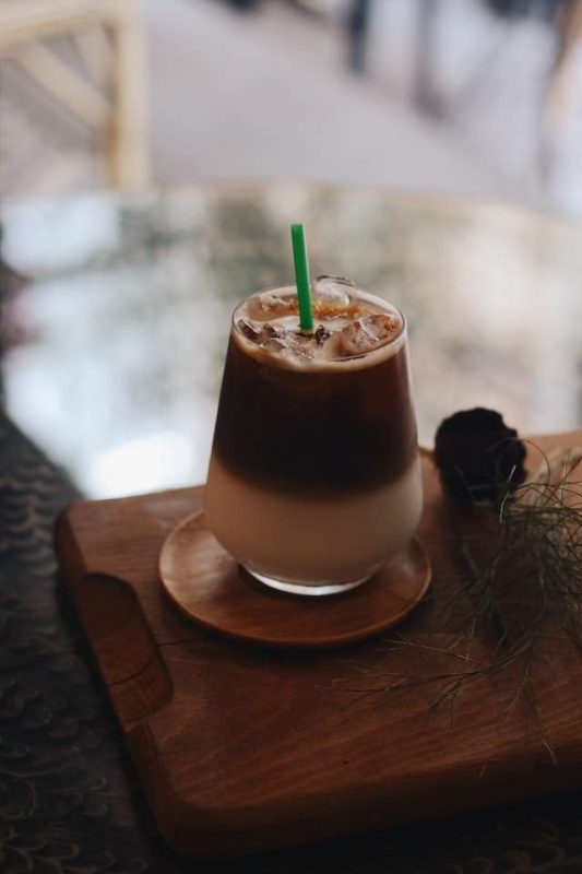 A glass of Irish cream iced coffee in a wooden saucer sitting on a table, a green straw in the glass highlights the color contrast.