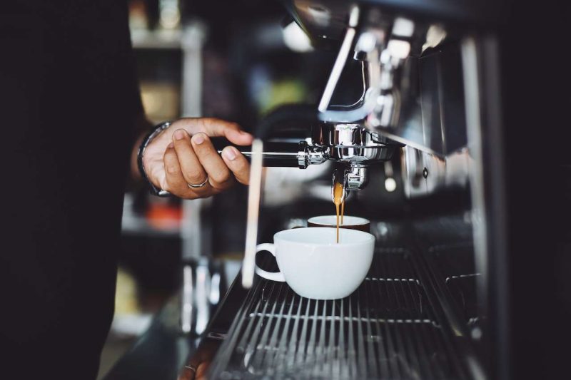 A man's left hand is on the handle of the espresso filter while the espresso machine is making the espresso and dripping into a white cup below. Coffee's caffeine is the main ingredient that coffee boosts your performance.