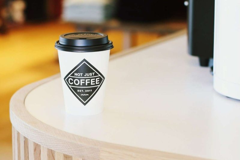 Screenshot of "Not Just Coffee" website home page, which shows a white to-go coffee cup with a black diamond shape company's logo with matching black cup lid. The cup is sitting on a white rounded corner counter edge. The background is out of focus. "Not Just Coffee" is one of the most popular coffee shops in charlotte nc.