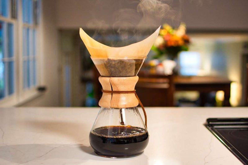 A pot of pour over coffee maker is brewing coffee with steam rising from it and the pot is 1/3 full. The pot is sitting on a white marble counter top next to a range, and against a blurry dining room background.