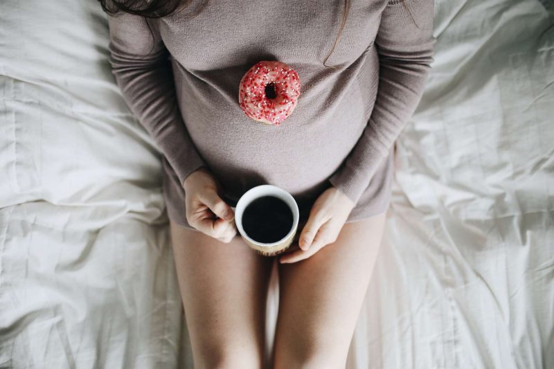 This picture shows a pregnant woman sitting in bed holding a cup of black coffee at the tip of her tummy which seems like she is almost due. She is wearing a long sleeve knitted shirt, her bear legs can be seen since the photo is taken from the top. There is a pink donut on top of her tummy about 6 inch higher to the coffee cup. Drink coffee while pregnant is general OK, but each individual has different reaction to coffee; so if it is unsure, please consult a doctor will be a good choice.