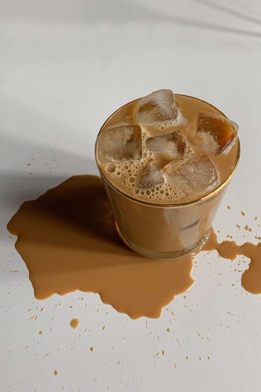 A glass of pumpkin spice iced coffee sits on a white table top and spills a bit on the table with a patch of coffee spill at the bottle of the glass. Use this image in Juan's iced coffee recipe.