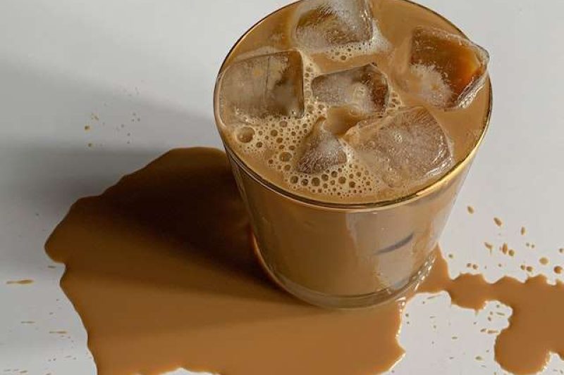 A glass of pumpkin spice iced coffee sits on a white table top and spills a bit on the table with a patch of coffee spill at the bottle of the glass. Use this image in Juan's iced coffee recipe.