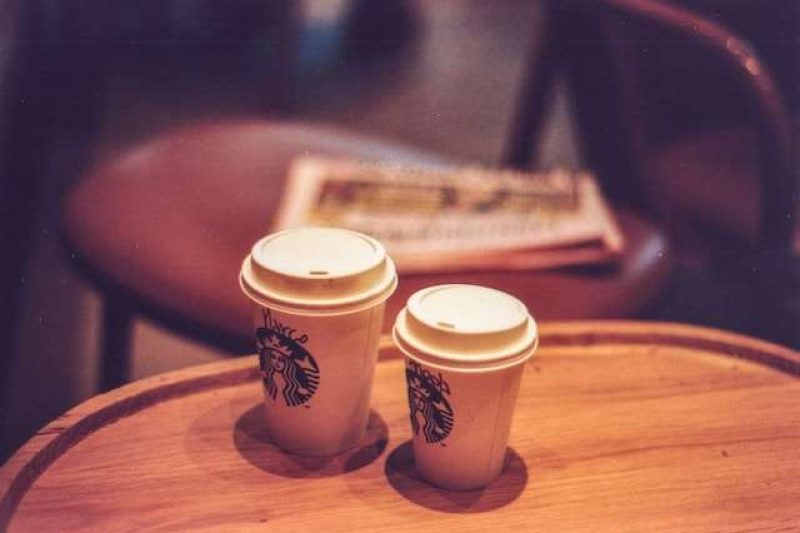 On a wooden oval coffee top sits two Starbucks to-go cups for hot coffee. The one slightly in the front of the picture is a short size, the one next to it, slightly in the back is a tall size. These are two of the Starbucks to-go cup sizes for its hot coffee.