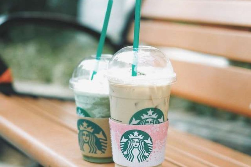 Two Starbucks To-Go Cups on a wooden park bench. Both cups show there is iced coffee in them through the cups semi transparent plastic. The tall one in the foreground is a "grande" size, the one next to it but in the background is a "tall" size. These are the 2 Starbucks To-Go cup sizes.