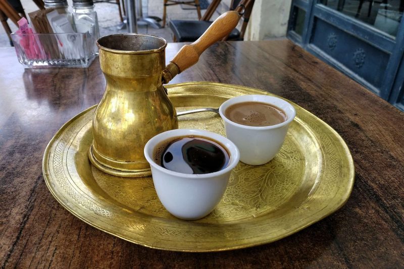 A brass Turkish coffee pot with wooden handle and two handle less small ceramic cups on a round brass tray on a dark wooden table. The cup in the foreground is a black Turkish coffee the other cup behind it is a white Turkish coffee. Part of a spoon can be seen between the coffee pot and the white coffee cup.