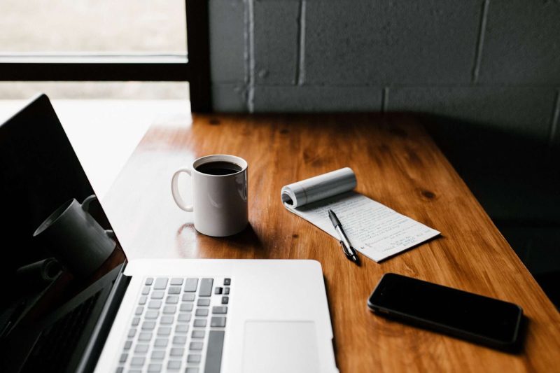 A laptop, a mug of black coffee, a notepad, a pen and a cellphone are on a nice grain wooden table which situates next to a window. The pen is on the notepad. People work while drinking coffee, is to take advantage of the effect that coffee enhances one's cognitive performance.